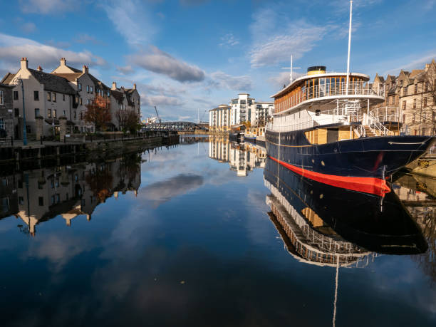 Still water and Autumn sunlight, with moored boats in Edinburgh’s historic Leith Harbour. Nearby old dock warehouse buildings have been converted into high end residential apartments, with views over the pretty scenic harbour, north east of the city centre. Reflections of the boats and buildings can be seen in the water.