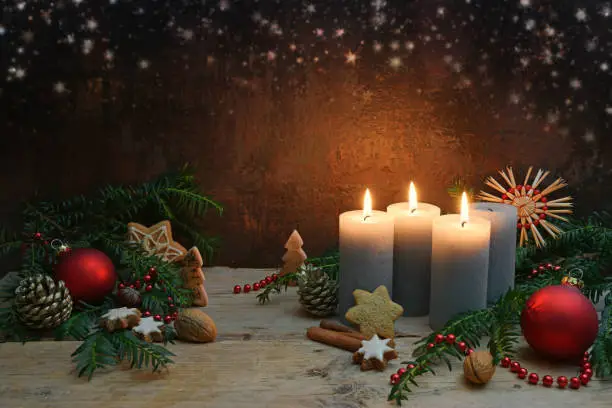 Third Advent, three of four candles are lighted, red baubles, branches and gingerbread cookies as Christmas decoration on rustic wooden planks, dark brown background with copy space, selected focus