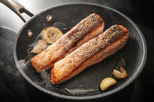 Two slices of fresh salmon fillet with a crispy fried skin in a black pan with garlic, thyme and lemon on the stove, cooking a delicious seafood meal, selected focus, narrow depth of field