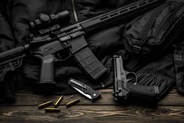 istock Modern automatic rifle with a telescopic sight on a dark background. Jacket, cap, gloves on a wooden table. The uniform of a guard or a mercenary. 1351567975