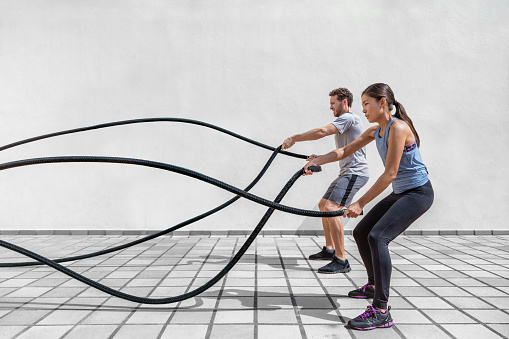 Fitness people exercising with battle ropes at gym. Woman and man couple training together doing battling rope workout working out arms and cardio for cross training exercises.
