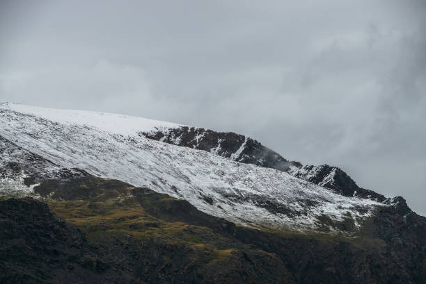 istock Minimalist highlands landscape with snow-capped mountains under cloudy sky. Snowbound high mountain range in overcast weather. Atmospheric minimalism with green mountains, white snow and gray sky. 1351573048