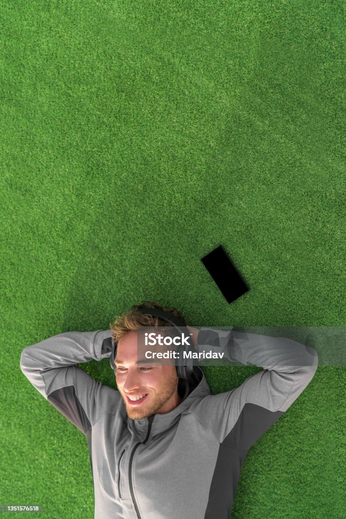 Listening to music for relaxation. Happy man lying down on grass enjoying podcast on mobile phone with wireless headphones. Summer lifestyle. Wellbeing Stock Photo