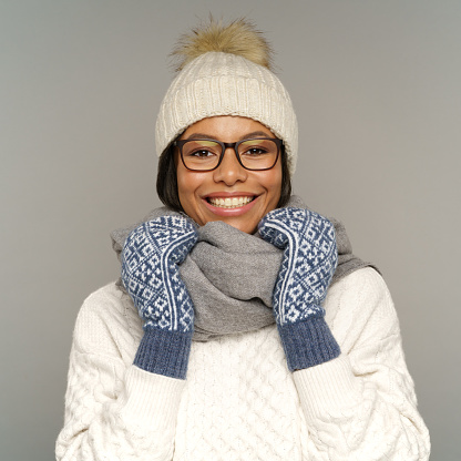 Warm winter clothes concept: happy smiling black female wearing knitted white sweater, hat, mittens and scarf protecting from cold weather and wintertime frost outdoors isolated over gray studio wall