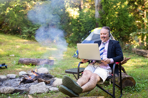 A man wearing only the top half of a suit and tie while he does an online video meeting with his laptop but actually he is enjoying the outdoors camping.