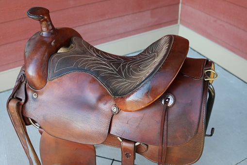 Old restored western roping saddle with shiny good quality leather on a saddle stand rack, with a view of the western seat and conchos and latigo offbillet.