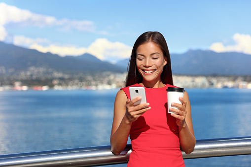 Businesswoman drinking coffee using mobile phone app in city to play video games or text sms online. Asian woman enjoying coffee break relaxing by watching smartphone content.