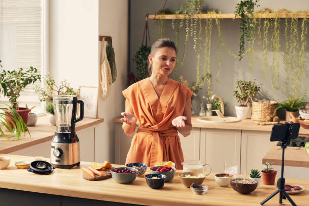 istock Young woman standing by kitchen table and cooking in front of smartphone camera 1351622010