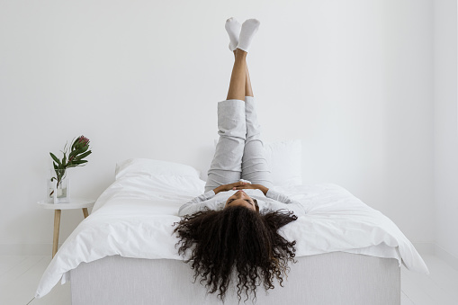 Happy and careless african american woman lying on bed in pajamas, raised legs up, resting at morning at home. Recreation, relaxation concept