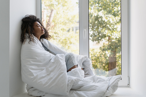 Serene afro american woman sitting on windowsill wrapped in blanket, relax, breathing fresh air at home. Peaceful young female warm up in bed cover, enjoy leisure weekend in hotel