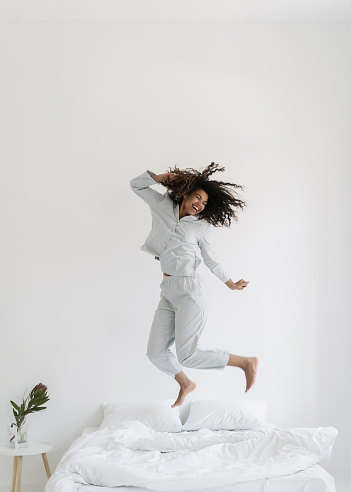 Vertical view of happy mixed race woman dancing in morning, jumping over bed in pajamas. Excited female spending day of in hotel or at home, having fun, feeling joyful mood. Careless, carefree concept