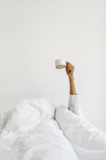 Concept of early morning wake up. Vertical view dark skinned woman in pajamas on bed, wrapped in warmth cozy blanket, raise hand with coffee cup. Female girl holding mug against white copy space wall