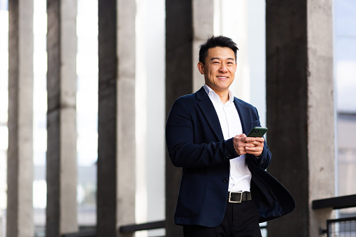 Asian businessman looking at camera smiling holding a mobile phone reading the news outside the office