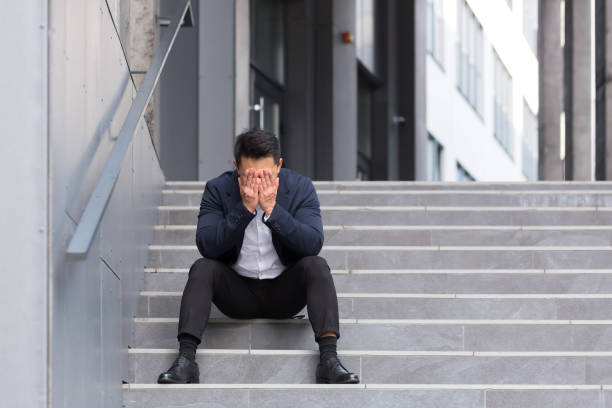 アジアのビジネスマンは、オフィスの近くの階段に座って深刻な頭痛を持っています - 敗北 ストックフォトと画像