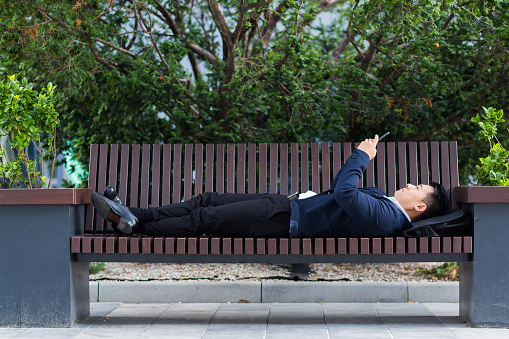 Male Asian businessman lying on a bench and resting after work uses a mobile phone to correspond and read the news