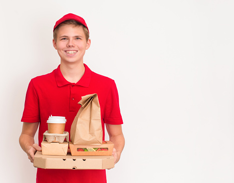 Smiling delivery boy giving food order and holding pizza box salad disposable paper cups and paper bag over white background.