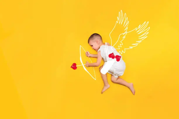 Happy toddler boy cupid in costume angel dress, wings, bows and hearts arrow isolated on yellow background. Valentines day, holidays, love concept. Copyspace. Selective focus.