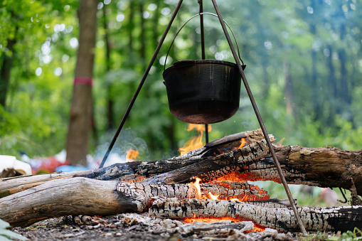 Camping outdoors. Cooking bowler hat hung on tripod over burning fire on the background of grass and chipped firewood