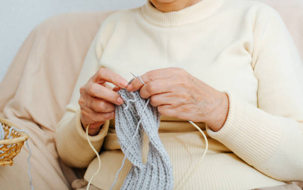 бабушка вяжет шарф, сидя на диване в помещении. крупный план руки пожилой женщины, держащей нити и иголки, занимающейся рукоделием, рукодели - grandmother knitting senior women senior adult стоковые фото и изображения