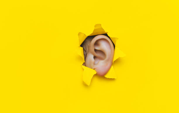 Close-up of a left woman's ear through a torn hole in yellow paper. The concept of eavesdropping, espionage, gossip, tabloids and the yellow press. Background with copy space. stock photo