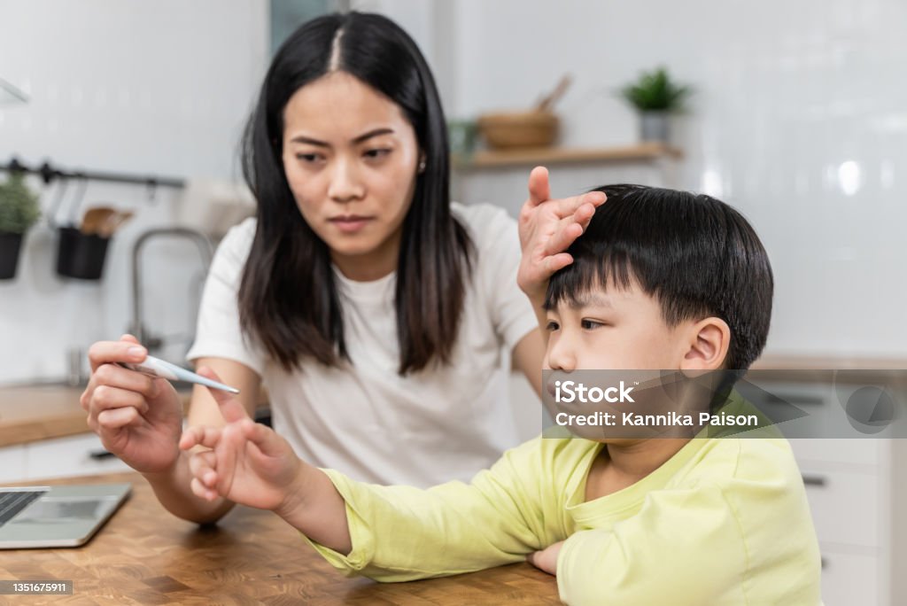 Young Asian mother measuring temperature of her ill kid with a thermometer. Portrait of Asian mother checking her sick sons temperature, looking at thermometer and touching forehead of her son. Respiratory syncytial virus Stock Photo