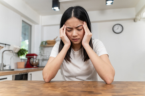 Stressed young Asian woman with headache while sitting alone in the kitchen at home. Exhausted female feeling headache and touching temples to relieve pain