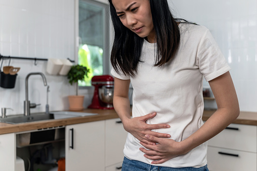 Young Asian woman suffering from stomachache or menstrual pain while standing in the kitchen at home.