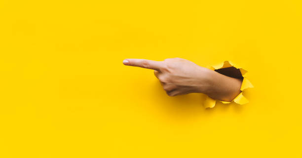 人差し指は左側を指します。黄色の背景。広告の場所。スペースをコピーします。女性の手が破れた紙の�穴に出てきた。 - 左利き ストックフォトと画像
