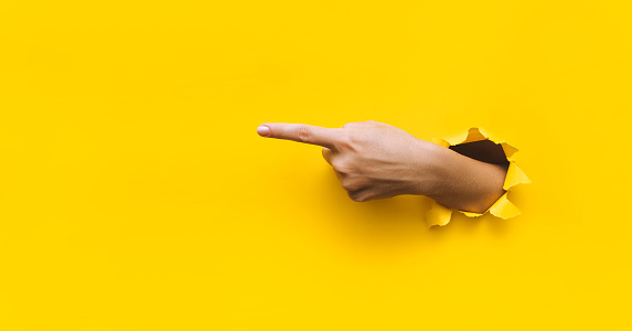 The forefinger points to the left side. Yellow background. Place for advertising. Copy space. The woman's hand came out into the torn paper hole.