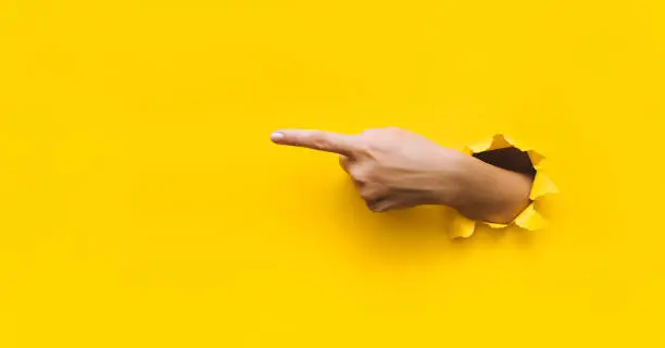 Photo of The forefinger points to the left side. Yellow background. Place for advertising. Copy space. The woman's hand came out into the torn paper hole.