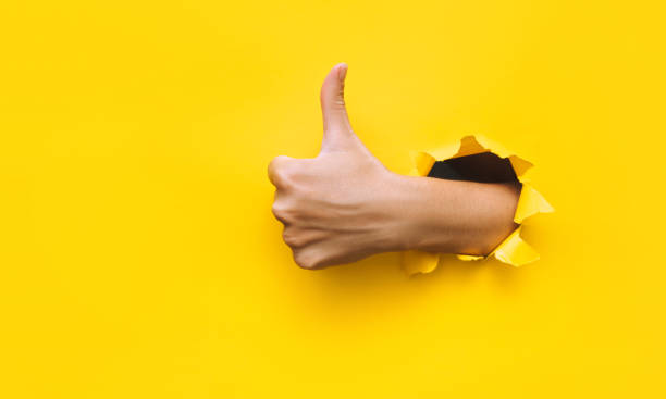 The female hand shows a thumb up gesture (like). Torn hole in yellow paper. The concept of positive attitude, approval and praise. Copy space. The female hand shows a thumb up gesture (like). Torn hole in yellow paper. The concept of positive attitude, approval and praise. Copy space. thumbs up stock pictures, royalty-free photos & images