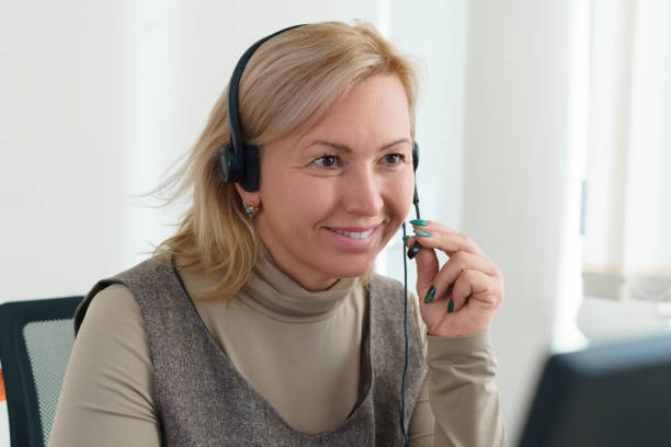 istock Smiling mature woman sitting in front of computer with headset speaking by online call while looking at camera. Home office. 1351679944