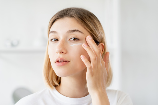 Under eye cream. Skincare moisturizing. Morning treatment. Pretty young woman applying beauty product on soft face skin at light bathroom.