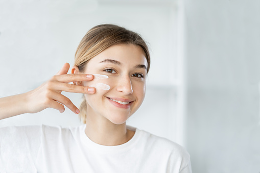 Home skincare. Moisturizing cosmetology. Facial treatment. Cheerful smiling woman applying organic cream on soft glowing face skin at light bathroom.