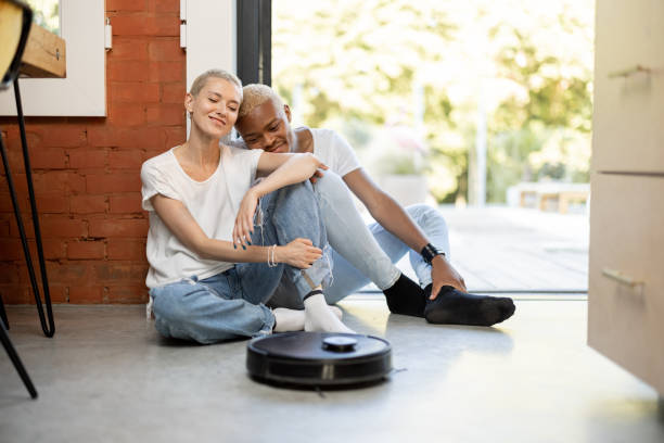 Couple sitting on floor with robot vacuum cleaner Multiracial couple sitting on floor at home with robot vacuum cleaner. European girl and black man spending time together. Concept of modern domestic lifestyle. Idea of house cleaning smart home family stock pictures, royalty-free photos & images