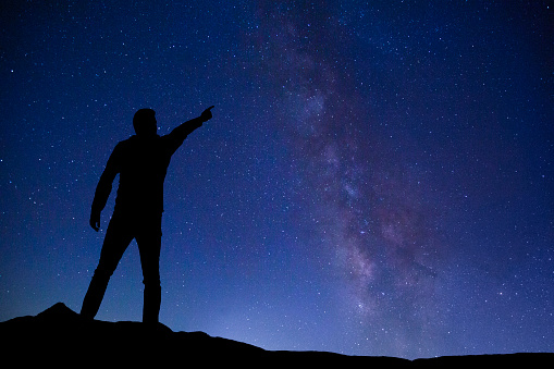 The silhouette of a man standing on top of a mountain, points to the Milky Way.