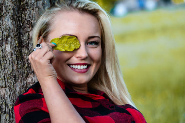 blonde girl leaning her head against a tree, playfully hides one eye behind a leaf, loving nature