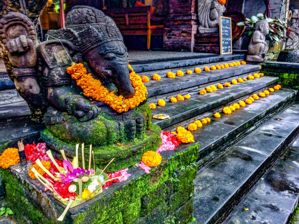 Horizontal closeup photo of a old carved stone statue of the Hindu mythological God, Ganesh, wearing a vibrant orange Marigold garland, with religious offerings around the base of the sculpture and flowers decorating the stone steps of a Cafe in Ubud, Bali