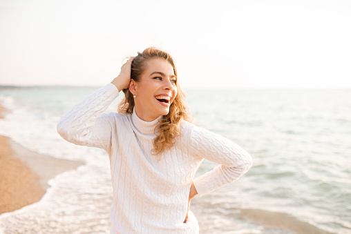 Laughing happy blonde woman wear white knit sweater resting over sea shore baat background. Summer season. Happiness. Cheerful adult girl.