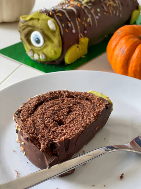 istock Close-up image of chocolate log cake slice on white plate with fork, Swiss roll covered in chocolate and designed to look like one-eyed monster, pumpkins, focus on foreground 1351727865