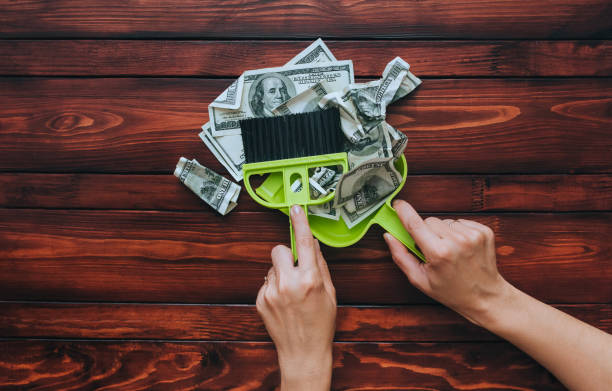 Female hands hold a brush and scoop and remove dollar bills from the wooden table. The concept of devaluation, depreciation, inflation and default, when money loses its value. stock photo