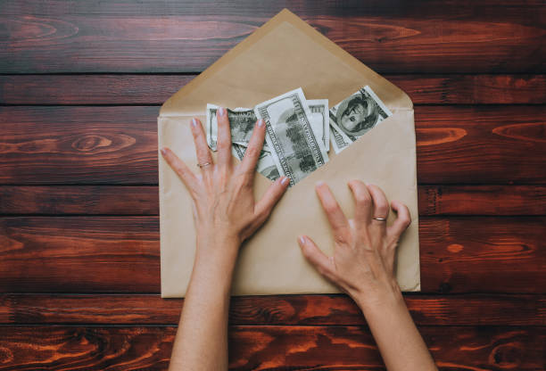 istock Two female hands, fingers spread out, hold an open beige craft envelope with dollars on a brown wooden background. The concept of donation, alimony, winnings and cashback. View from above. 1351733554