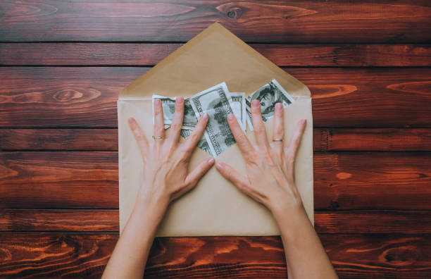 istock Female hands, fingers spread out, hold an open beige craft envelope with dollars on a brown wooden background. The concept of donation, alimony, winnings and cashback. View from above. 1351733557