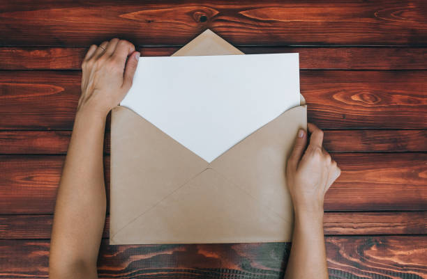 istock Young woman's hands opening a beige craft paper envelope with a blank white letterhead on a brown wooden table. Concept of letters, messages by mail. Copy space, mockup. 1351734643