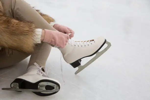 Closeup of female in fur coat and pink gloves wearing white skates on ice rink in winter day.Woman tying skates.Weekends activities outdoor in cold weather, sport and leisure concept