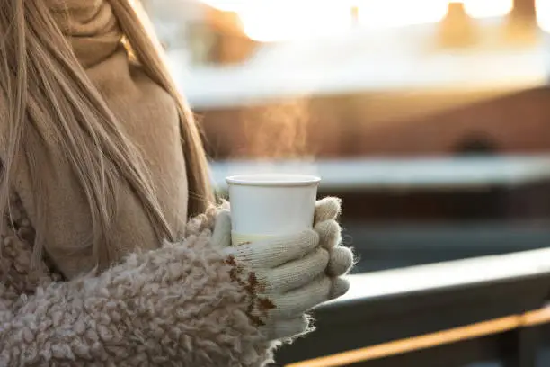 Female hands in white mittens holding steaming white cup of hot coffee or tea in cold winter sunny day, closeup.Winter time
