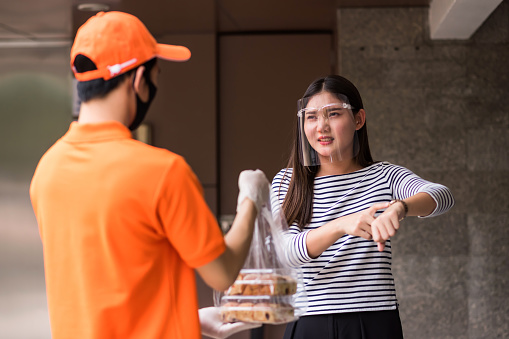 Angry hungry Asian woman with face shield show watch to complain  food delivery delay to courier deliveryman with mask  starving girl at office with covid-19 protect to prevent delta pandemic.