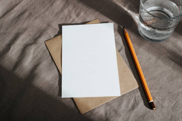 istock Greeting vertical white card with empty space for invitation, wishes mock-up. Craft envelope, blank card, pencil and glass of water on rustic vintage textile background with shadows 1351788681