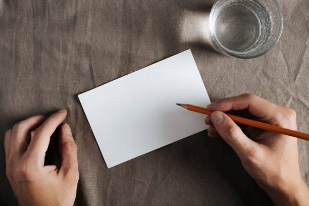 istock Top view greeting card mock up with empty space for text invitation, wishes or congratulations. Hand holding pencil, white blank card, glass of water on textile vintage background, flat lay 1351788694