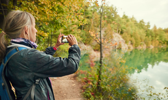 Senior woman in outdoor gear taking photos of a scenic lake while out for a hike in a forest in autumn